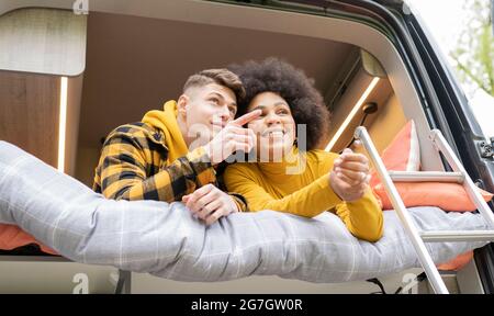 Low angle of African American Woman smiling with young boyfriend while lying down pointing away on bed in van during road trip looking away Stock Photo