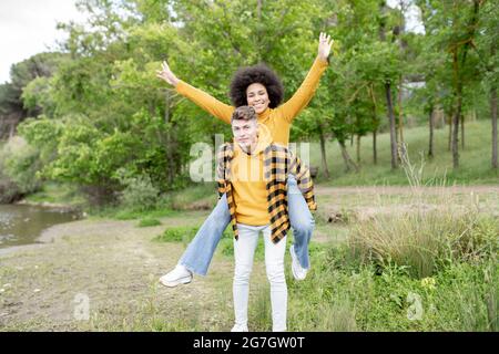 Young man giving piggyback ride to optimistic black girlfriend with raised arms while spending rime on coast of river in summer Stock Photo