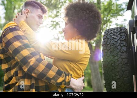 Low angle of young multiethnic man and Woman in casual clothes embracing each other while standing near car on sunny day in countryside Stock Photo