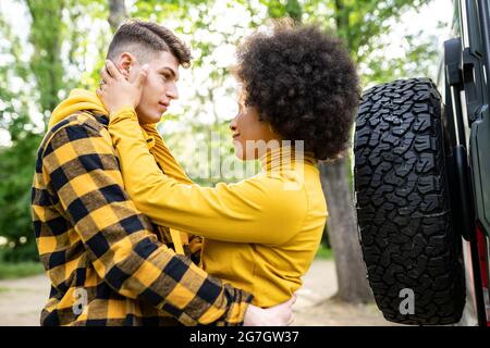 Low angle of young multiethnic man and Woman in casual clothes embracing each other while standing near car on sunny day in countryside Stock Photo