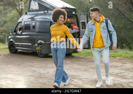 Diverse young man and Woman with longboard holding hands and walking on countryside road on summer weekend day near caravan Stock Photo