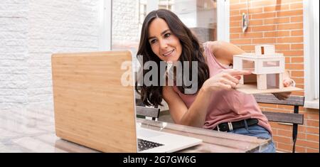 Smiling female architect showing model of house while talking on video chat via netbook and working on remote project Stock Photo