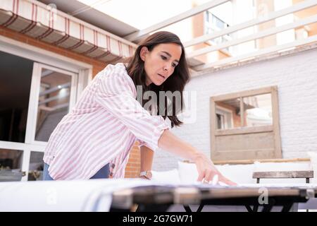 Positive female architect standing at table while analyzing and pointing at blueprint during work on project Stock Photo