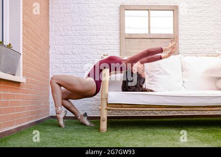 Side view of flexible graceful ballerina in pointe shoes and bodysuit balancing on tiptoes and showing backbend while leaning on sofa on terrace Stock Photo