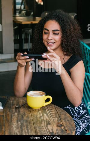Positive young Hispanic curly haired female taking photo of cup of fresh aromatic cappuccino while spending free time on cafe terrace Stock Photo