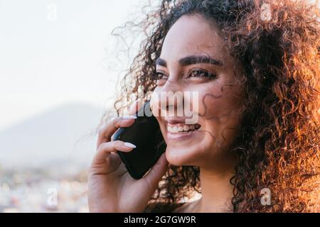 Pleased young Hispanic female with long curly hair talking on mobile phone and smiling happily on sunny summer evening outdoors Stock Photo