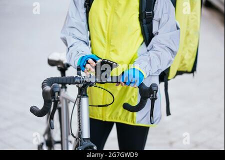 Crop view of anonymous young Woman with thermal bag browsing smartphone with GPS map on bicycle handlebar while delivering food on city street Stock Photo