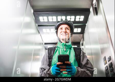 Low angle of glad female courier in uniform and helmet smiling and browsing cellphone in elevator while making delivery Stock Photo