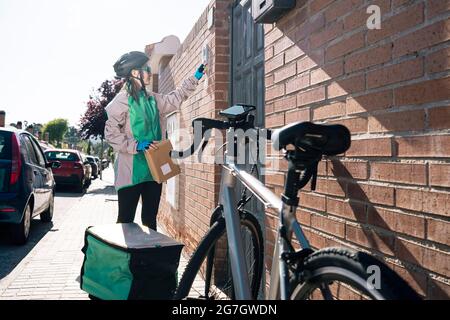 Female courier with parcel pressing button on intercom device on brick wall while making delivery on sunny day in town Stock Photo