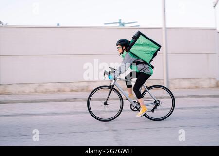 Side view of female courier with thermal bag riding bike on street road while delivering food in city, motion blur Stock Photo