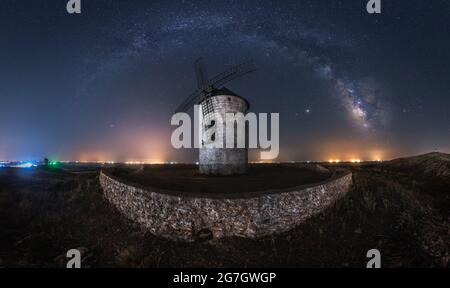 Picturesque scenery of Milky Way in dark night sky above aged stone windmill tower with glowing lights in distance Stock Photo