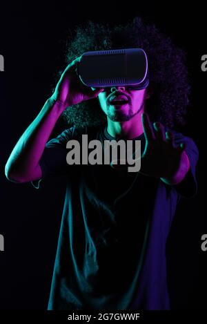 Excited Latin male with Afro hairstyle and in VR goggles experiencing virtual reality on black background in studio with neon lights Stock Photo