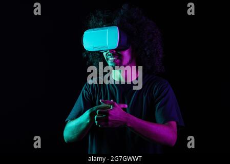 Excited Latin male with Afro hairstyle and in VR goggles experiencing virtual reality on black background in studio with neon lights Stock Photo
