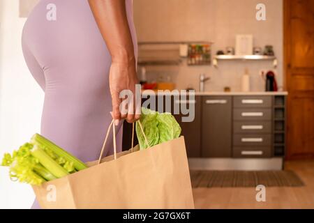 Side view of crop anonymous ethnic female with ripe green celery and lettuce in shopping bag standing in kitchen at home Stock Photo