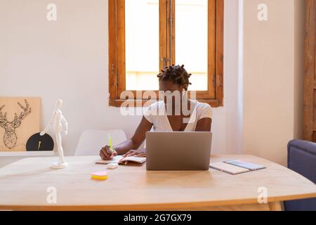 Focused African American female freelancer taking notes in notebook while working on project and sitting at table with laptop at home Stock Photo
