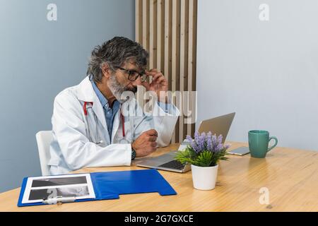 Focused elderly male doctor sitting at table in medical office while working with laptop in clinic Stock Photo