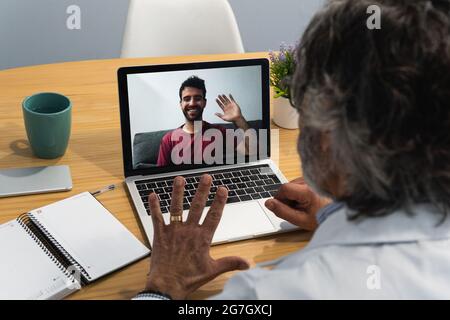 High angle of unrecognizable senior male doctor waving hand to patient while having video call during telemedicine appointment via laptop Stock Photo