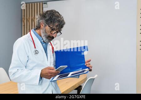 Focused elderly male medic in uniform and with x rays in folder standing in hospital and browsing mobile phone Stock Photo