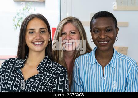 Delighted multiracial female colleagues looking at camera and smiling while sitting in workplace together Stock Photo