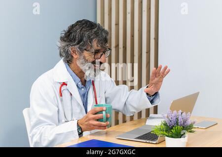 Smiling senior male doctor waving hand to patient while having video call during telemedicine appointment via laptop Stock Photo