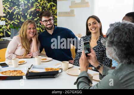 man taking photo of group of diverse coworkers sitting together at table during lunch in kitchen of office Stock Photo