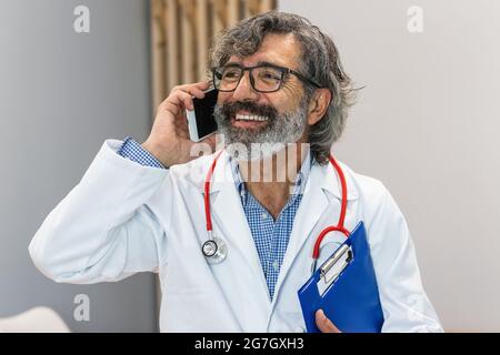 Cheerful elderly male medic in uniform holding folder standing in hospital while talking on mobile phone Stock Photo