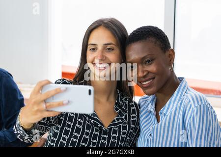 Cheerful multiracial female colleagues taking self portrait on smartphone while having lunch together in kitchen at workplace Stock Photo