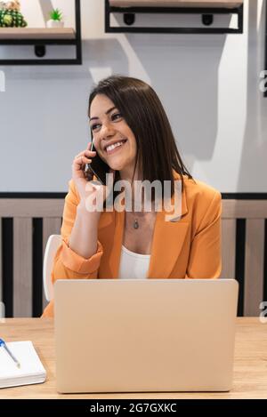 Smiling female freelancer sitting at table with laptop and using a phone Stock Photo