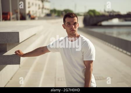 Handsome male in white t shirt standing on promenade in city on sunny day and looking at camera Stock Photo