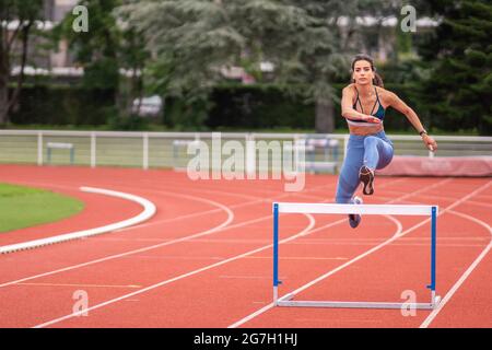 Full length of confident young Hispanic sportsWoman jumping over barrier while running on red track of sports stadium Stock Photo