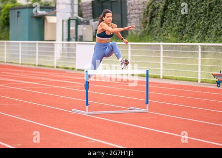 Full length of confident young Hispanic sportsWoman jumping over barrier while running on red track of sports stadium Stock Photo