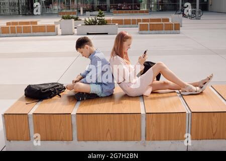 Portrait of cute young students wearing casual, sitting back to back, holding mobile devise in hands, seriously looking at screens, schoolyard near ca Stock Photo