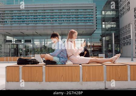 Portrait of cute young students wearing casual, sitting back to back, holding mobile devise in hands, seriously looking at screens, schoolyard near ca Stock Photo