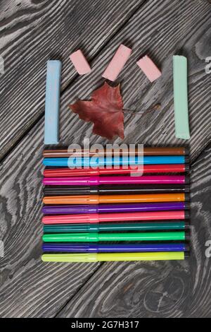 School accessories. Markers and crayons of different colors lie on pine boards. Nearby are fallen maple leaves. Stock Photo