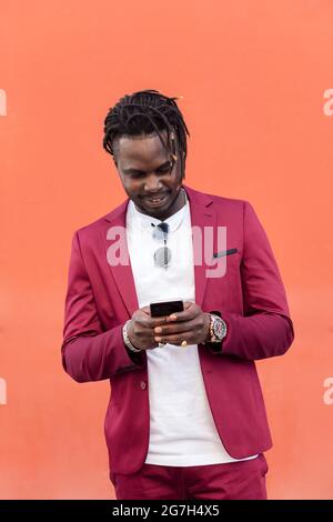 vertical photo of a stylish african businessman in suit typing a message on his smart phone in front of a red background, copy space for text, concept Stock Photo