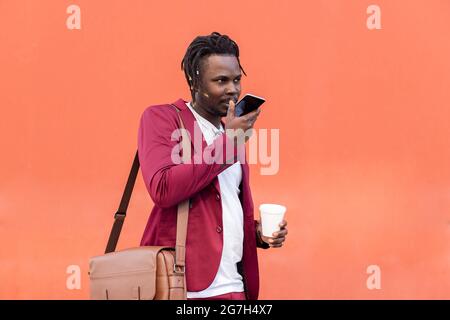 stylish black businessman dressed in suit with briefcase and coffee records a voice message on his smart phone in front of red background, copy space Stock Photo