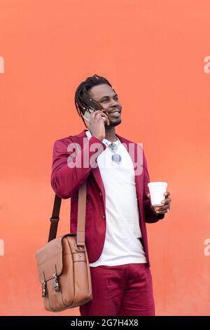 vertical photo of a stylish black businessman dressed in suit with briefcase and coffee talks on smart phone in front of red background, copy space fo Stock Photo