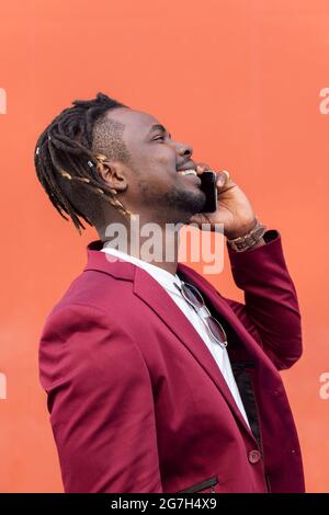 vertical profile portrait of a stylish african businessman in suit laughing and talking on phone in front of red background, copy space for text, conc Stock Photo