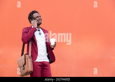 stylish african businessman dressed in suit with briefcase and coffee talks on smart phone in front of red background, copy space for text, concept of Stock Photo