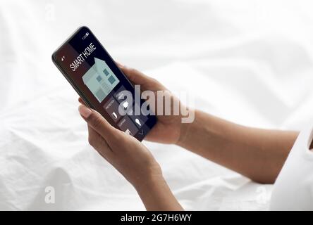 Smart home and mobile app for managing security, lighting, climate. Smart device for remote managing. Young woman lies in bed and controls functions o Stock Photo