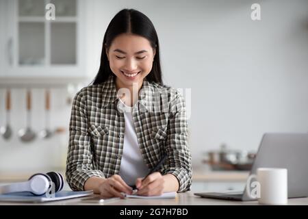 Positive young asian woman sitting at kitchen, taking notes while attending online class, using modern laptop, copy space. Happy chinese lady having o Stock Photo