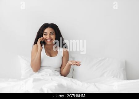 Great morning news. Happy african american lady in pajamas talking on smartphone in bed, empty space. Positive black woman making call on her mobile p Stock Photo