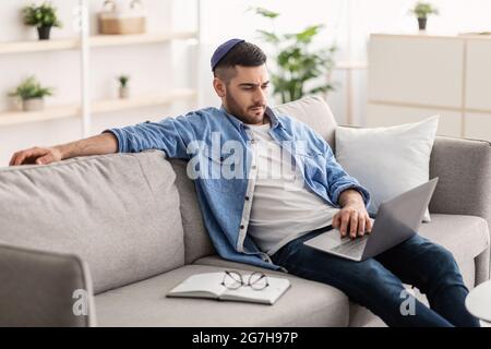 Portrait of focused young jewish guy wearing yarmulke using personal computer sitting on sofa in living room and working. Millennial man watching vide Stock Photo