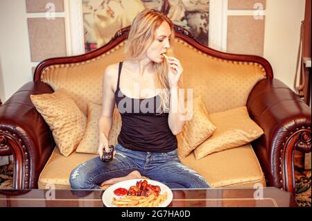 Beautiful young adult blond woman watching TV set and eats fried potato grilled chiken wings with ketchup tomato sauce Stock Photo