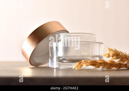 Open glass jar for empty face or body cream with golden lid on wooden table and oat spikes with beige isolated background Stock Photo