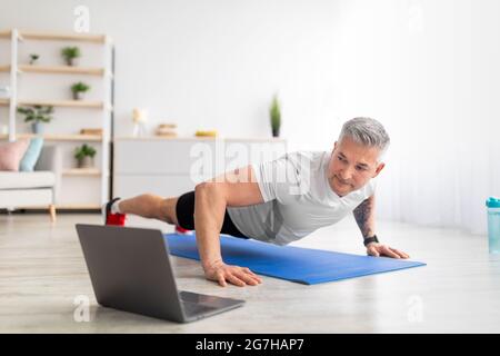 Fit mature man in sportswear doing push-ups, using laptop, having online fitness class from home, exercising in living room, copy space. Sporty man en Stock Photo