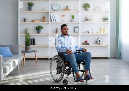 Full length of impaired African American guy suffering from disability depression, sitting in wheelchair at home. Young black man feeling sad and desp Stock Photo