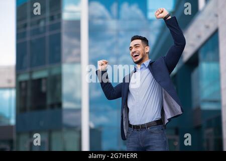 Emotional young entrepreneur middle-eastern guy in stylish outfit celebrating successful business deal, standing by office building, raising hands up Stock Photo