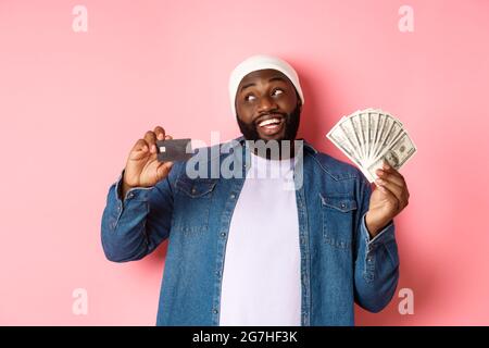 Dreamy african-american man showing credit card and dollars, thinking about shopping and smiling, standing over pink background Stock Photo