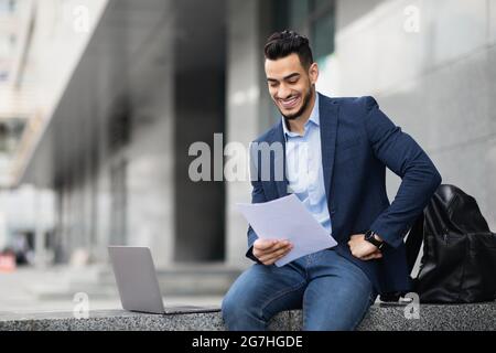 Young arab entrepreneur sitting by office building with laptop, holding documents and smiling, copy space. Confident cheerful guy in suit getting read Stock Photo
